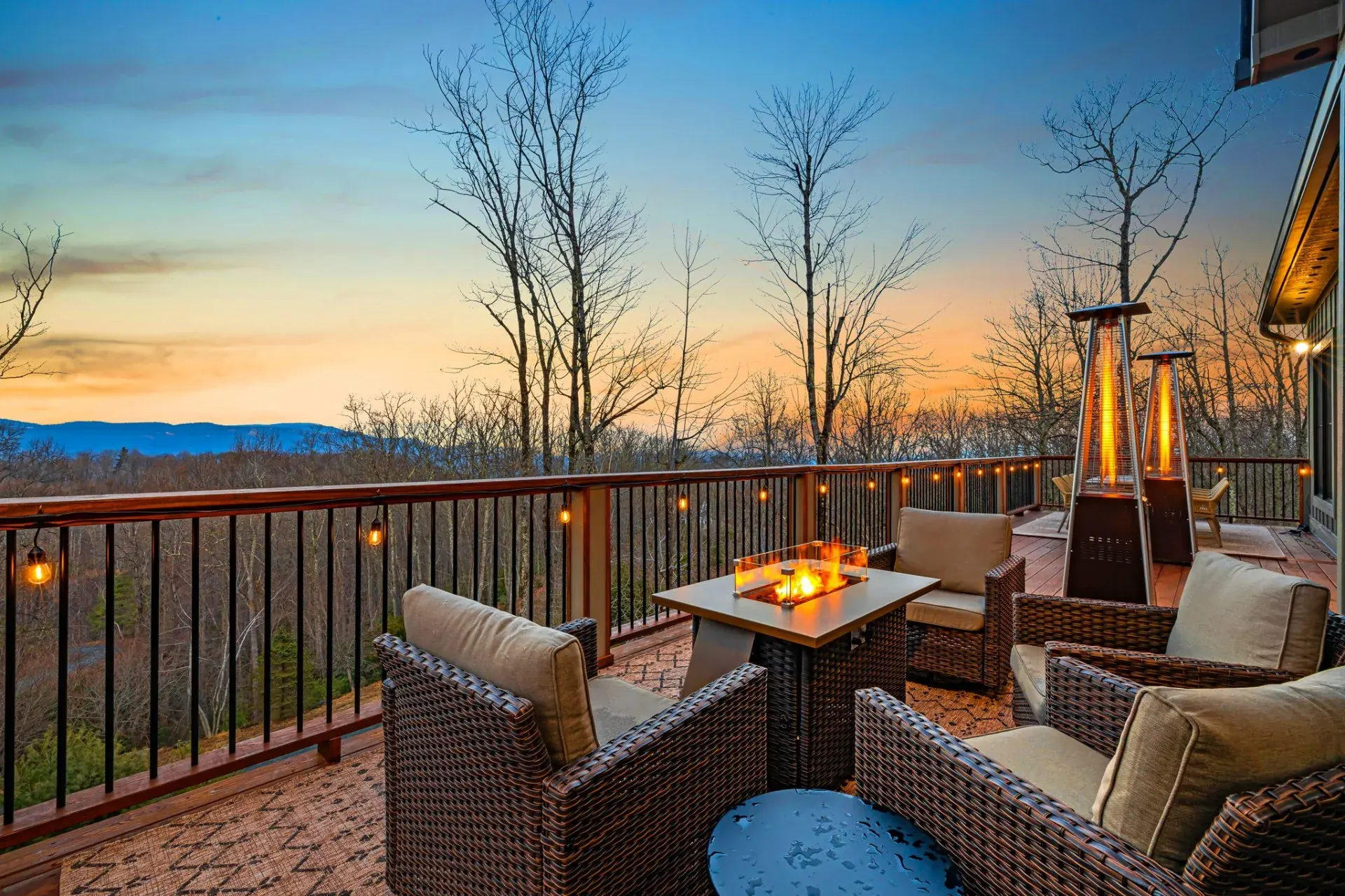 Deck lounge with a fire pit table, wicker chairs and string lights overlooking a mountain sunset