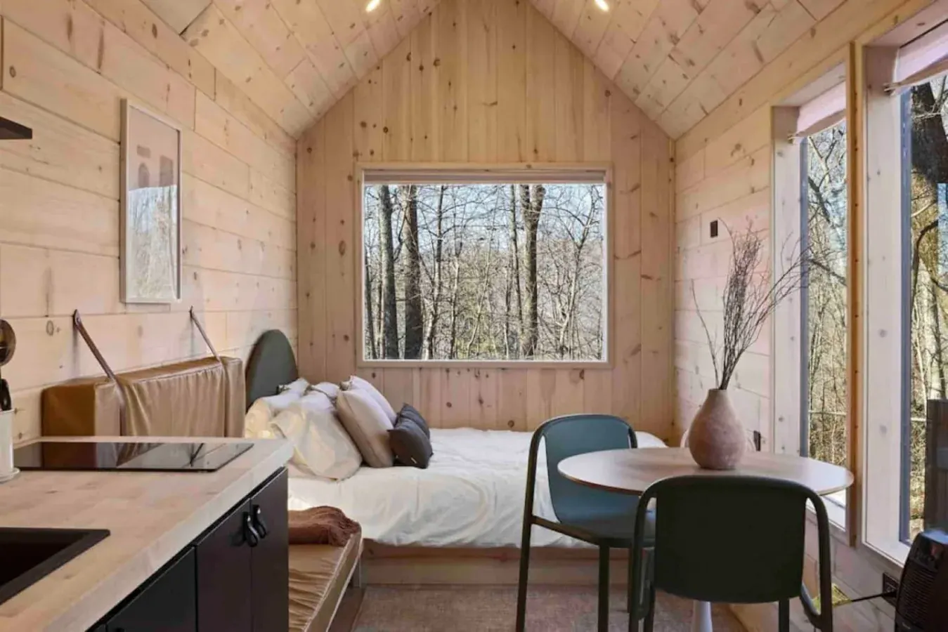 Tiny cabin loft with a vaulted wood ceiling, bed and dining table beside forest view windows